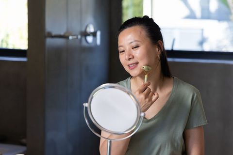 Mature asian woman using jade roller at vanity looking into mirror for skincare self care