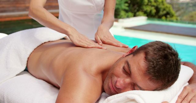 Man lying face down on a massage table covered with white towels. A therapist performs a back massage in an outdoor spa environment by the pool. Ideal for promotions related to wellness, relaxation, leisure retreats, and luxury spa services.