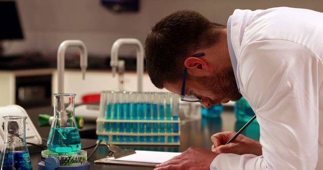 A Caucasian male scientist or lab technician is meticulously recording observations in a laboratory setting, with copy space. Surrounded by scientific equipment like test tubes and beakers, his concentration reflects the precision required in scientific research.