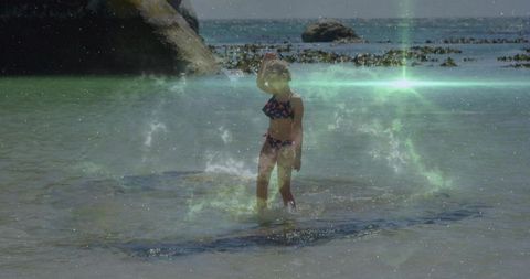 Child in floral swimsuit wading in ocean near rocky shore with added magical effect