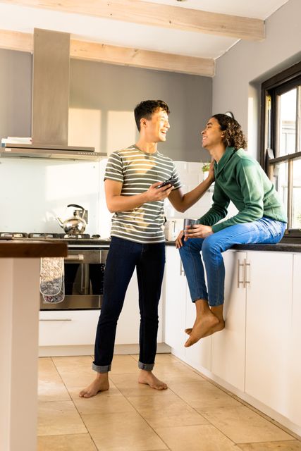 Multiracial cheerful man holding phone and looking at boyfriend holding coffee on kitchen counter. Gay couple, technology, drink, copy space, unaltered, love, together, homosexual, lifestyle, home.