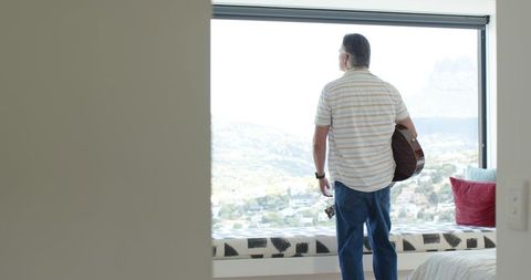Man holding guitar gazing out large window at panoramic valley view in modern bedroom
