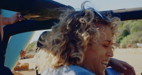 Curly blonde woman laughing in jeep while friend carries surfboard on sunny beach drive