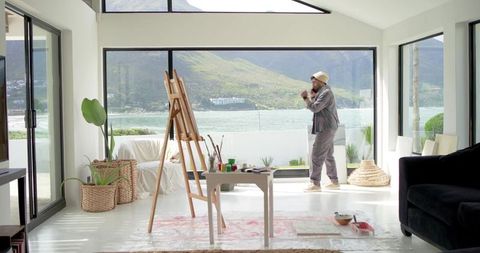 Indian artist painting by waterfront in bright modern studio showing ocean view and easel