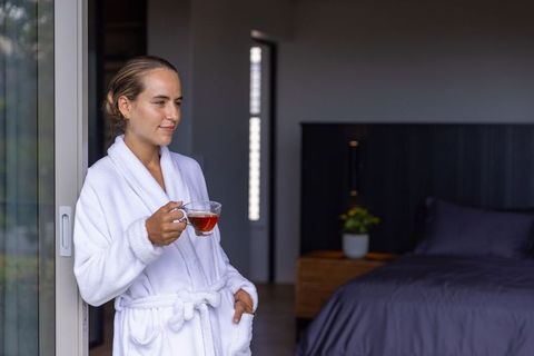 Woman in white bathrobe holding glass teacup at sliding door enjoying morning calm
