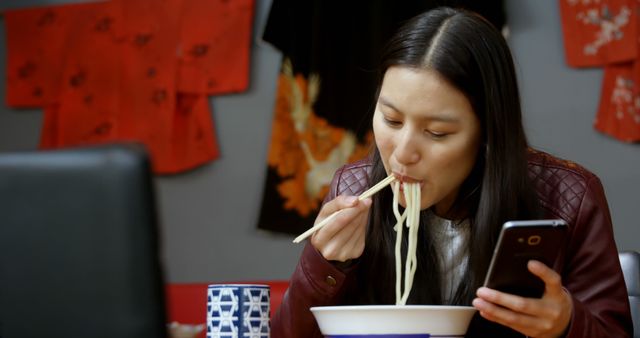 Young woman eating a bowl of noodles with chopsticks while looking at her phone, set in a traditional Japanese environment. This image can be used for content about modern dining habits, cultural experiences, or technology's role in daily life.