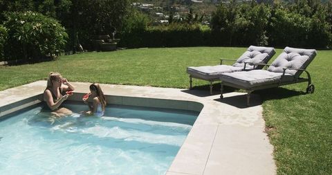 Mother and daughter enjoying watermelon by backyard pool on sunny summer day