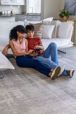 Diverse couple sharing smartphone while relaxing on modern living room rug