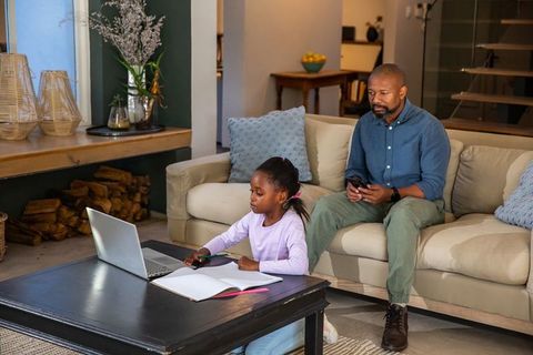 African american father supervising daughter studying on laptop in cozy living room