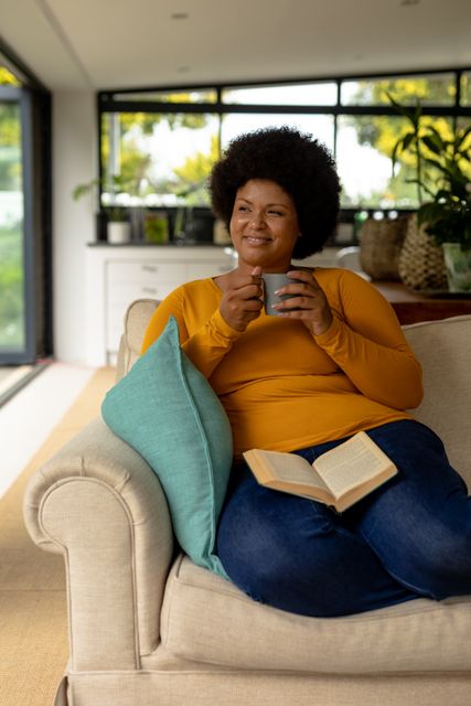 This image depicts a smiling African American woman enjoying a moment of relaxation at home with a coffee cup and a book. Ideal for use in lifestyle blogs, articles about leisure activities, advertisements for home comfort products, or promotions for coffee brands. It conveys a sense of comfort, contentment, and the simple pleasures of domestic life.