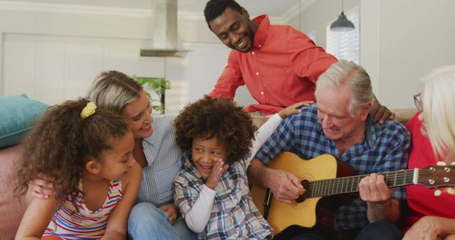 Image of diverse family siting on the couch and grandfather playing the guitar. Family life, spending time together with family.