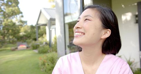 Smiling Korean woman gazing upward in sunny backyard with modern patio and garden