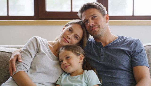 Parents cuddling daughter on sofa under wood-framed window, relaxing family moment