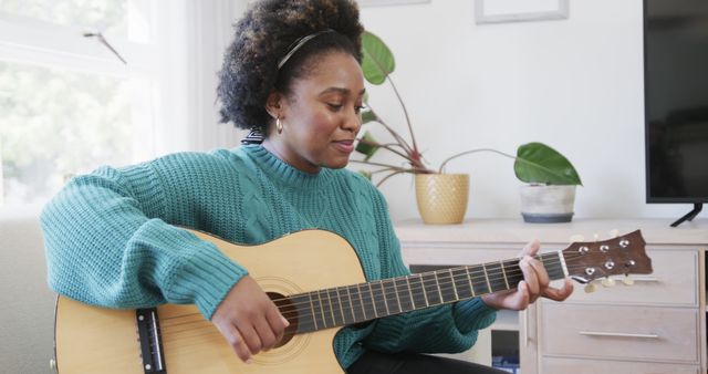 Image depicts a woman sitting in a cozy living room, strumming an acoustic guitar. She wears a teal sweater and seems focused and content. There is a potted plant and wooden furniture in the background. This could be used for articles or posts related to relaxation, hobbies, home lifestyle, creative pursuits, music learning, or promoting a balanced lifestyle.