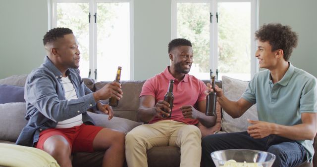 Three young adult men are sitting on a sofa, laughing and drinking beer together in a bright living room. They are holding beer bottles and engaging in a friendly conversation. This image can be used for content related to friendship, social gatherings, leisure activities, and promoting relaxing environments or alcoholic beverages.