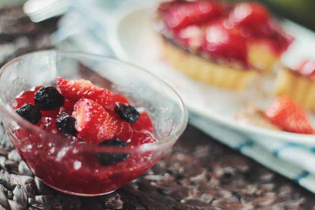 This close-up shot shows a colorful and fresh berry jelly dessert inside a glass bowl. Strawberries and blackberries are combined in a vibrant jelly mixture. In the background, there is another plate of dessert perfectly complementing the glass bowl's contents. This image is ideal for food blogs, recipe websites, nutrition articles, and social media posts focused on healthy eating, homemade recipes, and desserts.