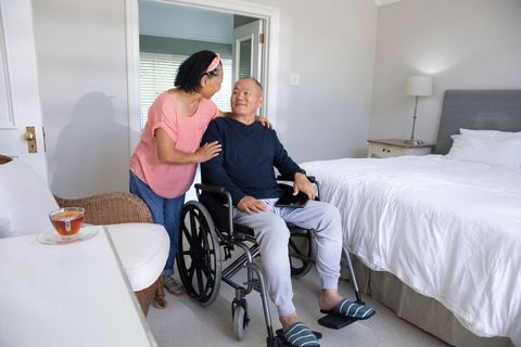 Asian senior couple sharing moment as man in wheelchair using tablet in cozy bedroom