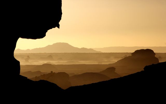 Silhouetted rock formations against a stunning desert landscape at sunset, with light haze creating an ethereal effect. Ideal for articles and publications about nature, travel, and adventure. Perfect for backgrounds, storytelling visuals, and desktop wallpapers.