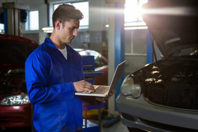 Mechanic in blue overalls using laptop for diagnostics in auto repair garage. Ideal for illustrating modern automotive repair, technology in workshops, and professional car maintenance services.