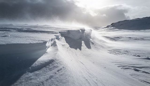 Wind-sculpted arctic cornice on snowy plateau with dramatic backlit sky and blowing snow