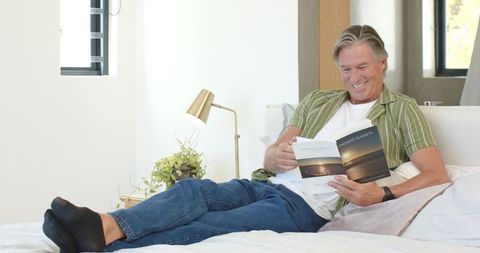 Mature man relaxing on bed reading paperback in bright minimalist bedroom