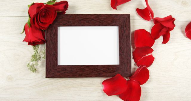 A wooden picture frame is surrounded by red rose petals and a bouquet of roses on a white wooden background, with copy space. Ideal for Valentine's Day, anniversaries, or romantic occasions, the frame offers space for a personal photo or message.