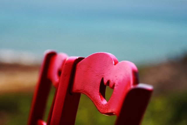 This image features a close-up view of a red wooden chair with a heart-shaped design, set against a blurred seascape background. Perfect for use in romantic-themed projects, coastal or summer promotional materials, and decor ideas that aim to evoke feelings of love and calmness. The vibrant red color of the chair adds a touch of warmth and passion.