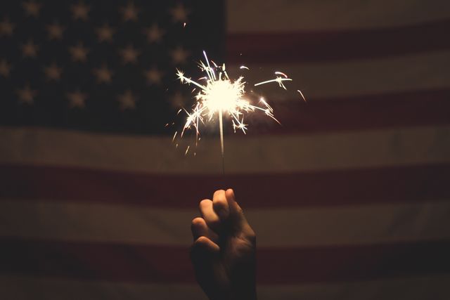 Hand holding sparkler in front of background with American flag, capturing the spirit of celebration and patriotism. This image is perfect for illustrating the Fourth of July, Independence Day, Memorial Day, or any event related to American pride and culture. Useful for websites, blogs, and publications focusing on holidays, national pride, or festive events.