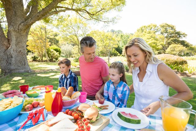 A family with parents and children enjoying a picnic in a park on a sunny day. They are surrounded by a variety of food including sandwiches, fruit, and drinks. This image can be used for promoting family activities, outdoor recreation, healthy living, and summertime fun.