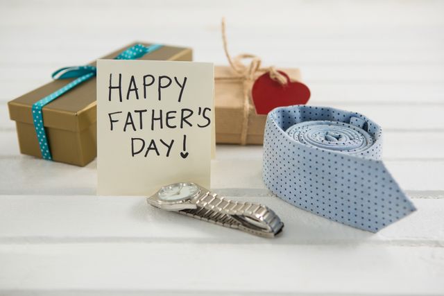 This image shows a Father's Day greeting card placed next to gifts on a wooden table. The gifts include a wrapped present, a tie, and a watch, symbolizing love and appreciation for fathers. Ideal for use in Father's Day promotions, greeting cards, social media posts, and advertisements celebrating the holiday.
