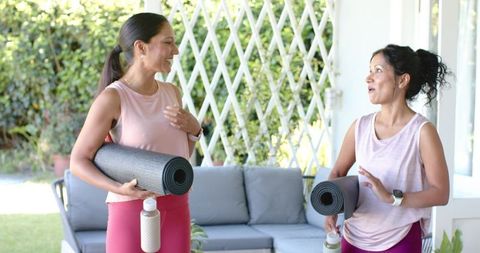 Chatting women holding yoga mats and water bottles on backyard patio wearing magenta leggings