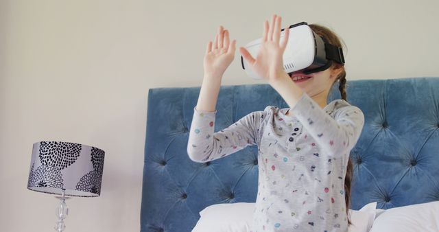 This shows a young girl wearing a VR headset, demonstrating the enjoyment of virtual reality technology. She stands in a bedroom with blue headboard, gray patterned lamp on bedside table adds context to the domestic setting. The joy and wonder on her face as she interacts with the virtual environment can be used in content about modern gaming, family tech experiences, staying home activities for kids, or the future of technology.