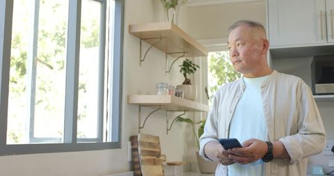 Senior Asian man using smartphone while gazing through sunny modern kitchen window