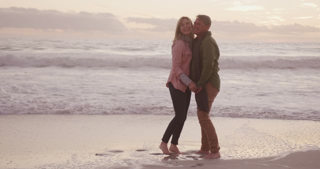 This image shows a couple holding hands and smiling while standing on a sandy beach with gentle waves in the background. The setting sun casts a warm glow, creating a romantic and serene atmosphere. Perfect for use in travel brochures, romantic getaway promotions, relationship counseling flyers, wedding invitations, or lifestyle blogs.