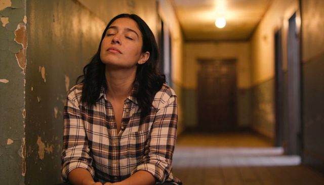 Young woman resting in dim peeling-hallway, plaid shirt, contemplative solitary portrait