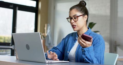 Asian woman working from home on laptop and smartphone during virtual meeting with glasses