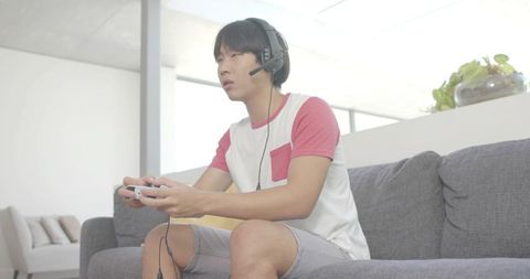Young man gaming on couch wearing headset and holding wired controller, focused player