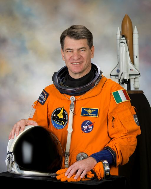 An astronaut dressed in an orange spacesuit representing the European Space Agency stands smiling with a helmet in hand, posed beside a model shuttle. Perfect for use in educational materials, promotional content related to space and science, and articles about space exploration and institutions like NASA.