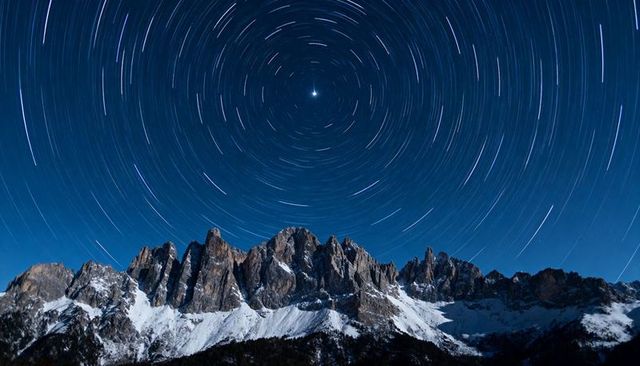 Circling star trails over snowy jagged mountain ridge under moonlit alpine night