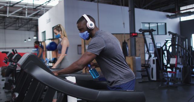 Fit african american man wearing face mask and headphones cleaning treadmill machine with disinfectant in the gym. social distancing quarantine lockdown during coronavirus pandemic