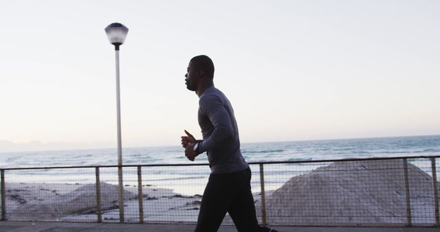 African American man jogging along a beachside path against a backdrop of the ocean and sunrise. Great for promoting a healthy lifestyle, outdoor fitness routines, activewear brands, and morning exercise benefits.