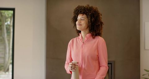 Woman in pink zip-up holding insulated bottle standing relaxed in sunlit living room