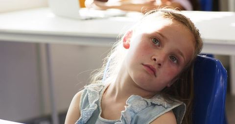 Thoughtful schoolgirl gazing upward while sitting at desk in sunlit classroom bathed in soft light