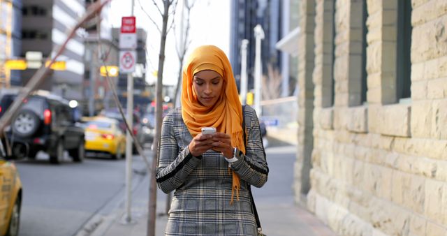 Woman wearing an orange hijab and a plaid dress walking on an urban street, texting on smartphone. Suitable for themes related to urban lifestyle, communication, technology use, and representations of Muslim women in modern city environments.