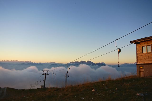 Photograph features a mountain cable lift silhouetted against a glowing sunrise above a sea of clouds. The fog blankets valleys below, creating a stunning landscape. Ideal for travel brochures, scenic backgrounds, and inspirational posters.
