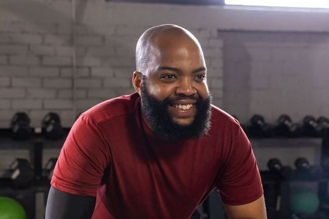 Smiling african american man leaning on gym equipment wearing maroon shirt for fitness motivation