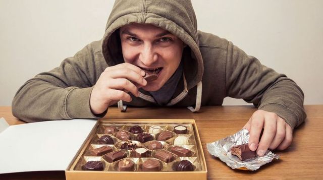 Hooded man sneaking chocolates at wooden table with open gold box and foil wrapper