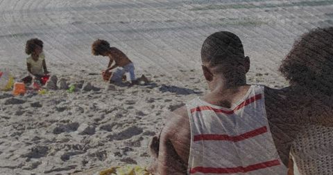 Father watching children building sandcastles on sunny beach during relaxed family vacation