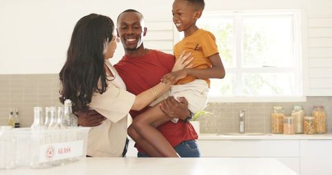 Laughing multiracial family embracing in sunlit kitchen, father lifting son, cozy bonding moment