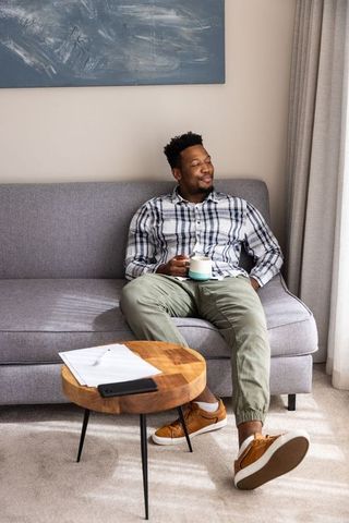 Serene african american man relaxing on gray sofa holding mug in sunlit living room